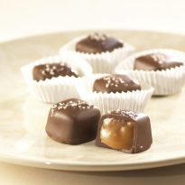 Milk Chocolate Sea Salt Caramels sit on white plate. Four (4) Milk Chocolate Sea Salt Caramels sit in white paper cups. Two (2) sit on the plate with one (1) cup open to reveal the soft caramel inside.