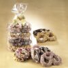 Assorted three (3) ring pretzels with various toppings, including rainbow seeds, white chocolate drizzle, rainbow sprinkles, and mini peanut butter cups. Seven gourmet pretzels lie on a brown background with seven (7) pretzels wrapped in a cello bag wrapped in a gold bow.